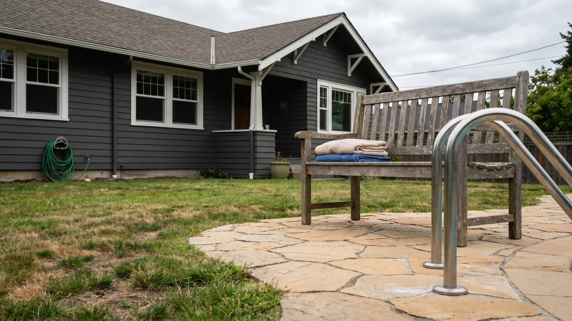 Small paver pad with a towel bench at the base of a pool ladder by a gray craftsman house