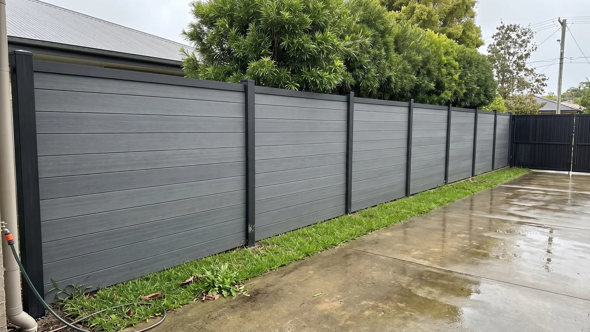 Mid-grey composite fence boards with black posts and cap along a wet concrete driveway