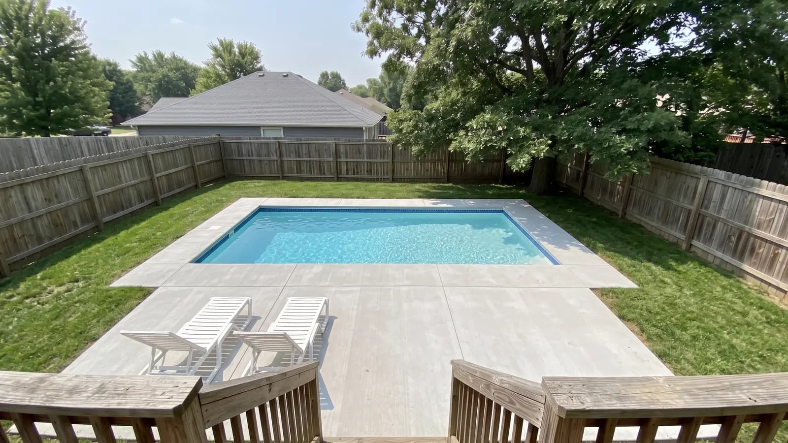 A rectangular inground pool with light blue water and a gray concrete deck in a fenced suburban backyard with an oak tree