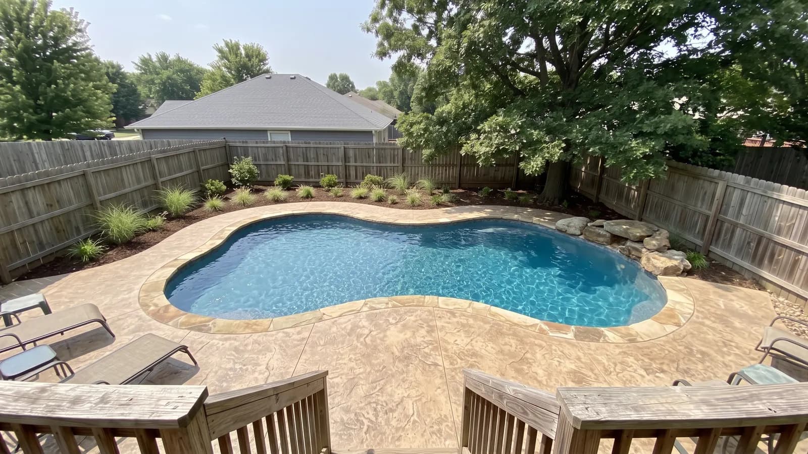 A freeform inground pool with soft curves, flagstone coping and boulders near an oak tree in a fenced backyard