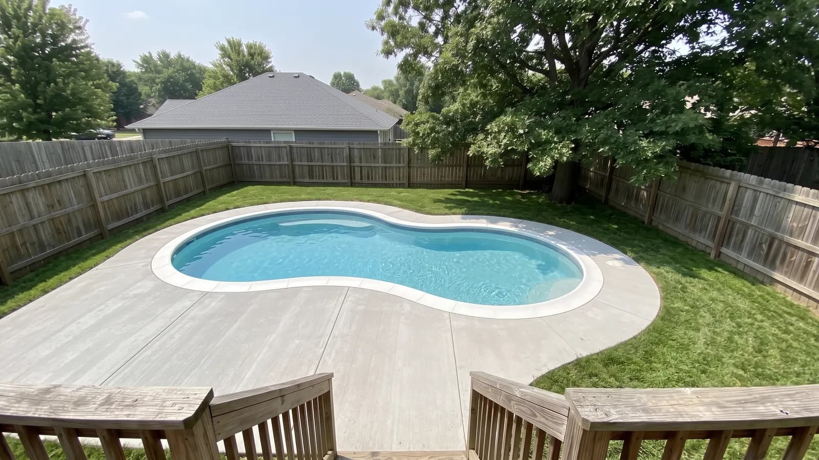 A kidney-shaped inground pool with white bullnose coping set into a wide concrete deck in a fenced backyard