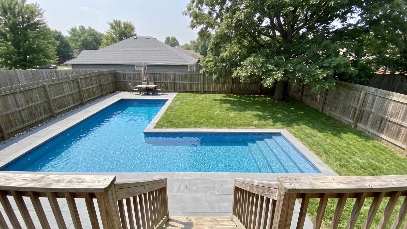 An L-shaped inground pool with a long arm across the yard, a shorter arm running toward the back fence and lawn inside the L
