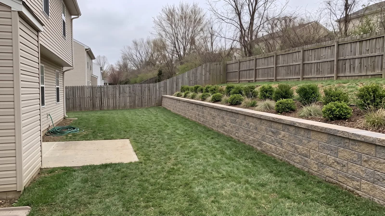 AI concept of a three-foot tan segmental concrete block retaining wall with a smooth cap holding back the backyard slope, with boxwood and grasses planted on top