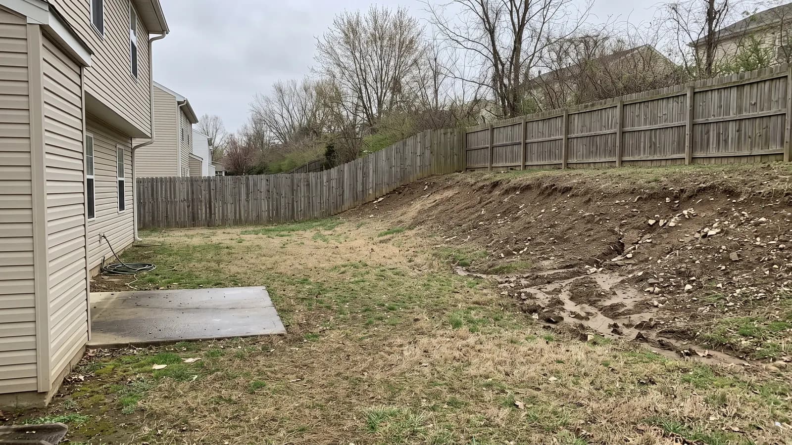 Retaining wall cost starts with a slope like this one: a bare, eroding grass bank beside a small concrete patio in an ordinary suburban backyard